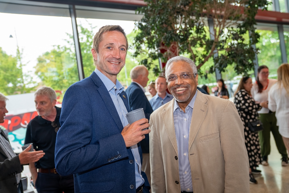 two smartly dressed men, one holding a reusable coffee cup, smiling to camera at an indoor event