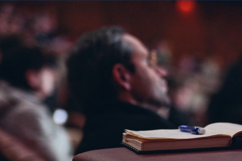 close up of an open notebook and pen on a table with blurred conference attendees in the background