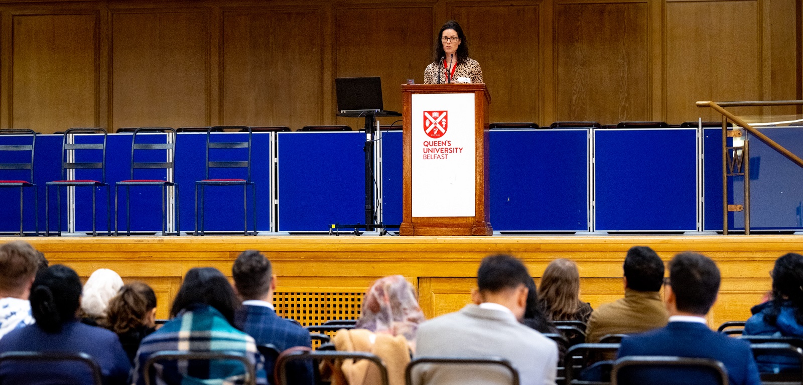 female postdoc/researcher presenting on stage to attendees at Queen's Postdoc Showcase 2023 in the Whitla Hall