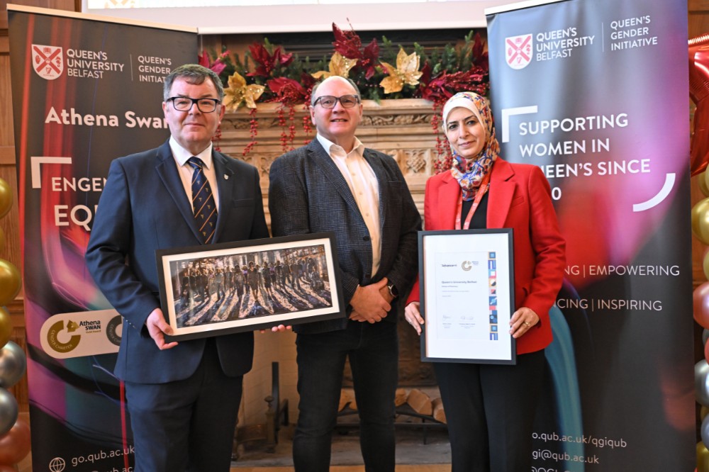 two men wearing glasses and suits - one holding a painting - and a woman holding a framed award certificate in an old event hall with large decorated fireplace, pop-up stands and presentation screen in the background