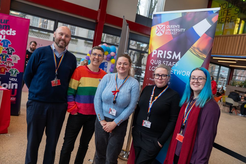 Five staff members from the PRISM LGBTQ+ network stand together smiling in front of a colourful PRISM banner.