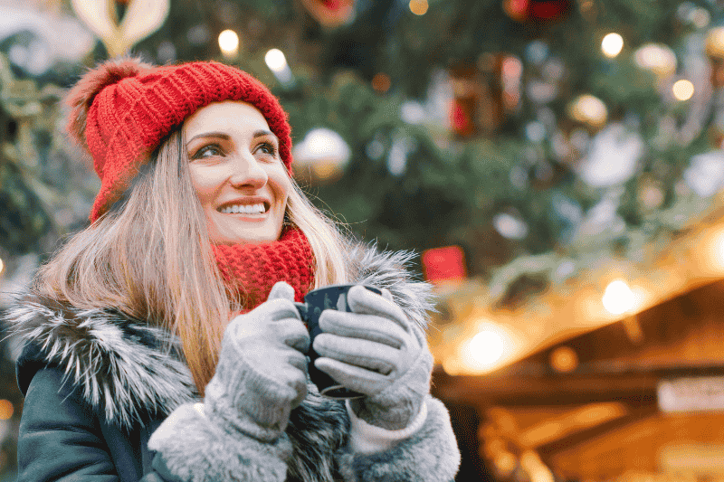 smiling young woman wearing warm winter clothes and holding a drink mug outdoors with a festive scene in the background