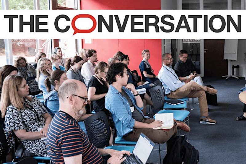 people with notebooks and laptops sitting in a large meeting room and listening to an information session. Image includes banner logo of 'The Conversation' news source.