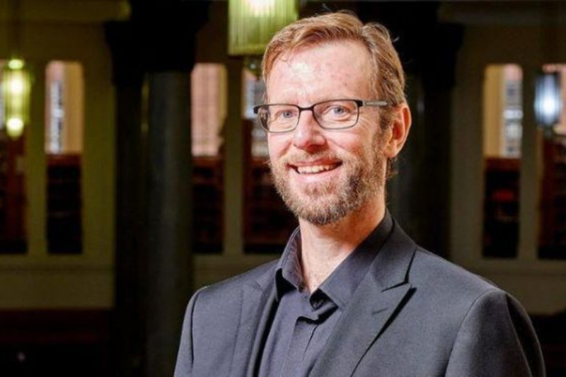 smiling man wearing suit and glasses looking to camera in an indoor setting
