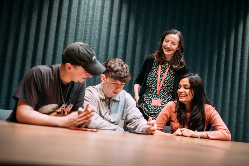 three young adults sitting at a table with a smiling female supervisor standing behind wearing a red lanyard