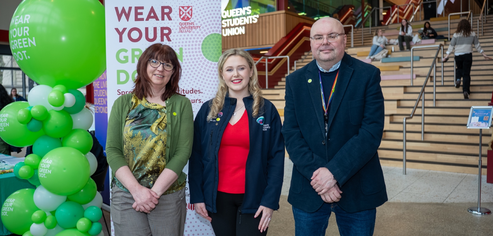 Two women and a man stand beside a poster and balloons reading 'wear your green dot' as part of a student safety campaign