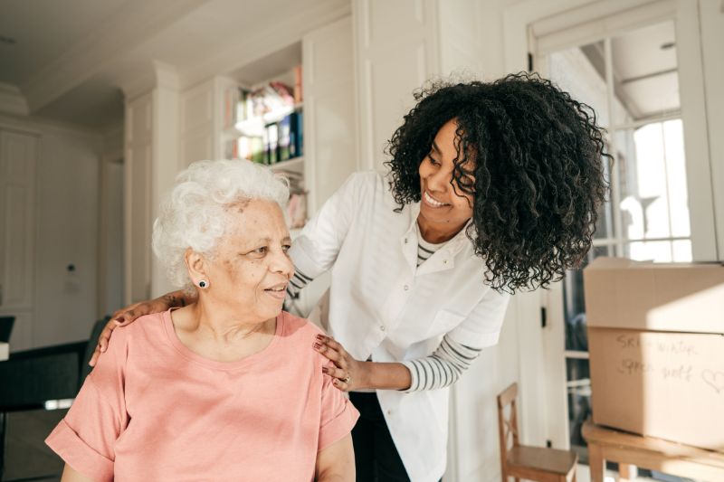 smiling, elderly woman seated indoors with younger woman speaking to and taking care of her