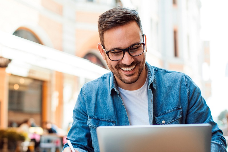 smiling man using a laptop and writing at an outside cafe space