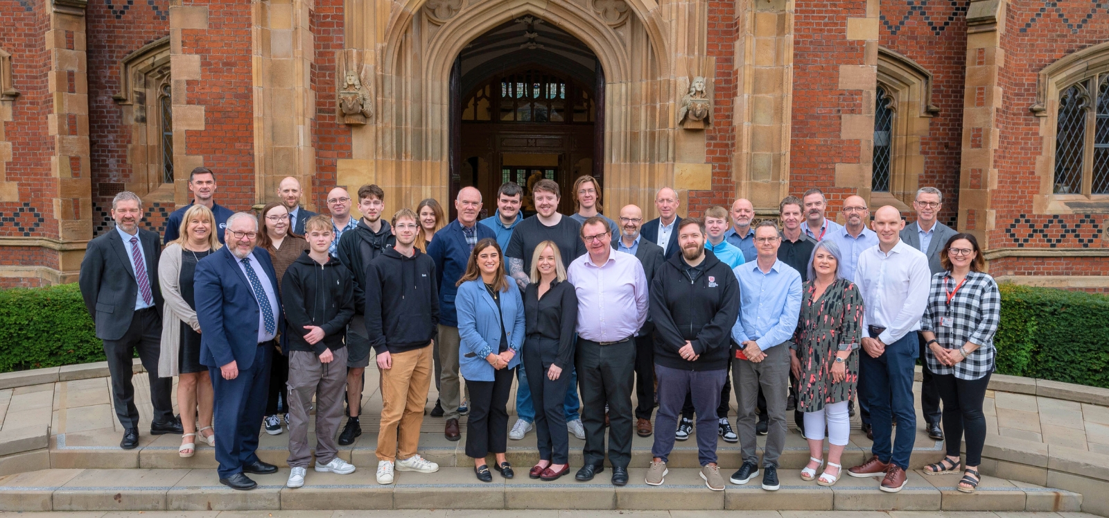 large group of smiling people standing in three rows on steps at the entrance to an old redbrick building