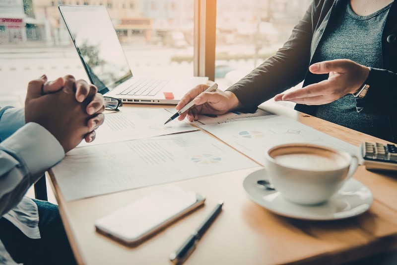 two people chatting over documents at a table - one person has a laptop and a cup of coffee