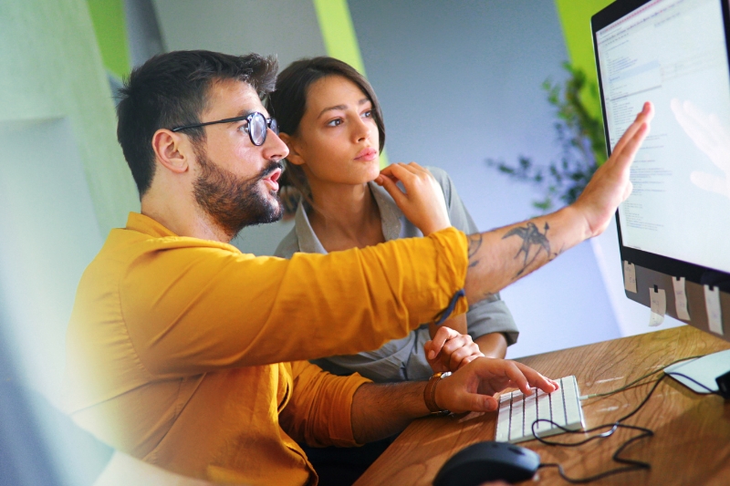 young man wearing glasses seated at an office desk, showing and explaining something on a computer screen to a young woman