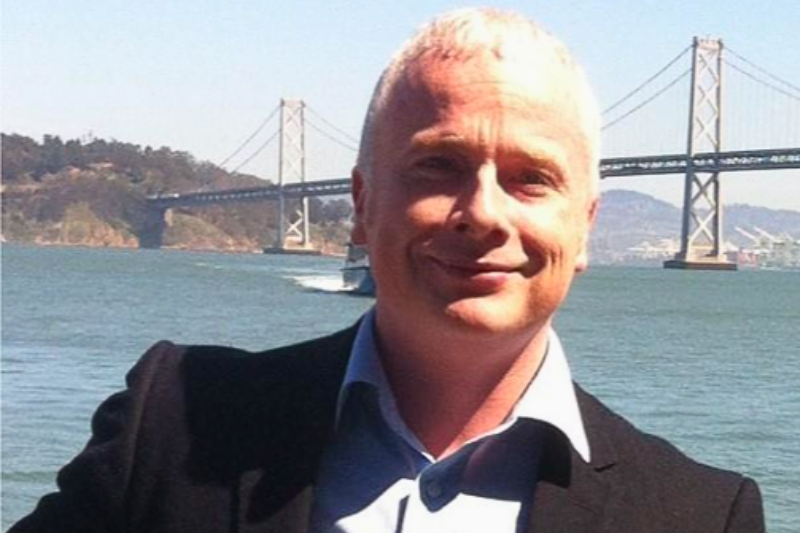 man in black jacket smiling with body of water and bridge in the background