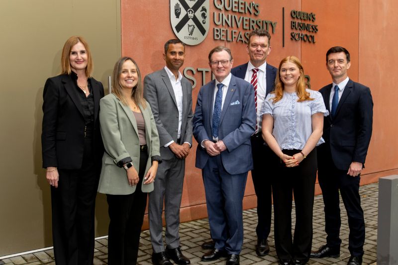 three women and four men in smart business clothing standing and smiling for the camera outside Queen's Business School, Queen's University Belfast