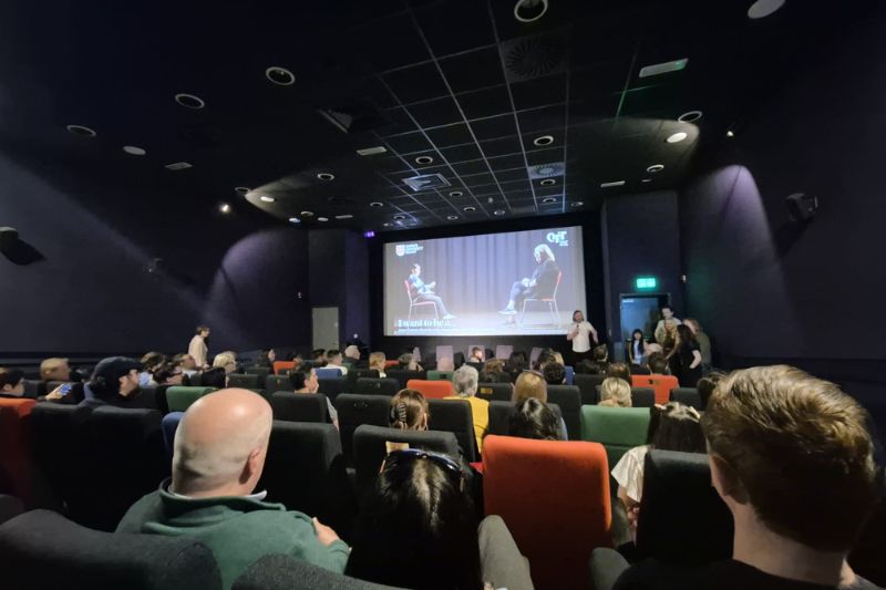 view from the back of an independent cinema screen-room showing a seated audience listening to a man standing and talking in front of the screen