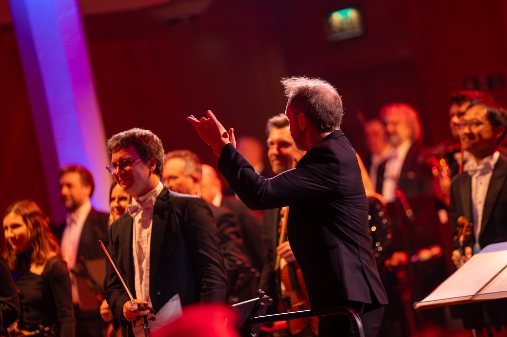 a conductor applauding while smiling orchestral players stand in the background