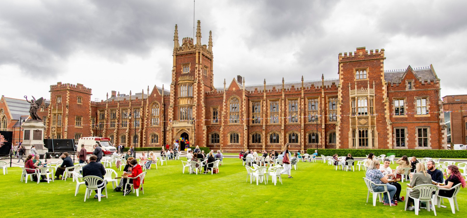 staff and students seated on the Queen's University Belfast front lawn at Queen's Pride Picnic 2024, with food trucks to the side and coloured bunting and balloons in sight