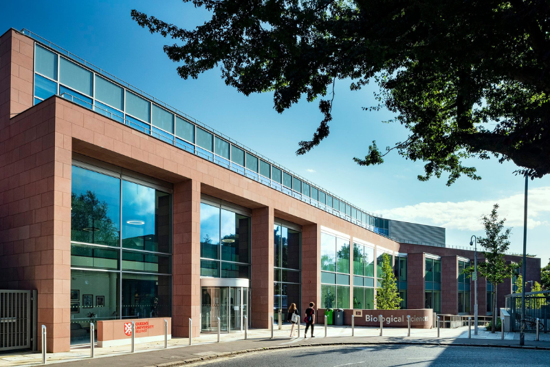 front entrance to a modern concrete and glass-fronted building on the corner of a road, pictured in sunny weather with blue skies and a shady tree on the right foreground. Wording on one wall reads 'Biological Sciences'.
