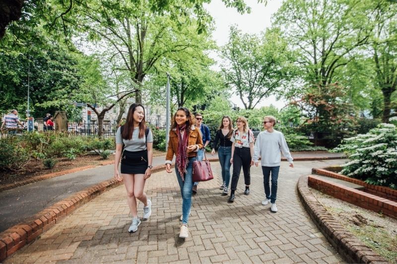 group of young people - students - walking and chatting on a pedestrianised pathway with trees towering behind