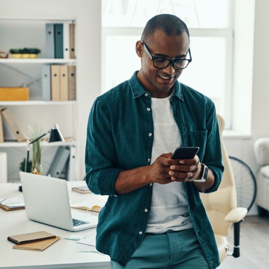 smart, smiling black man perched on desk using mobile phone in bright home office environment