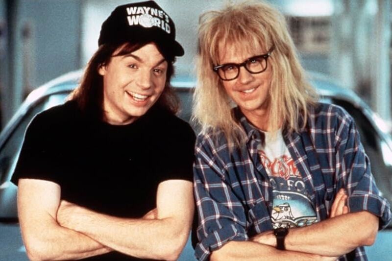 Wayne and Garth, the two main characters in the film Wayne's World, smiling to camera. One dressed in black t-shirt and black hat with 'Wayne's World' writing on it; the other with scruffy hair, wearing thick-rimmed glasses and lumberjack checked shirt