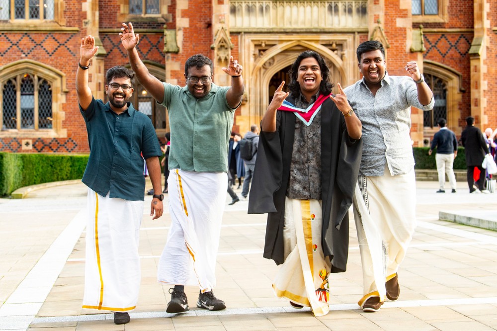 four young men of South Asian origin, partly wearing traditional dress, walking in a celebratory way towards the camera with an old redbrick building in the background